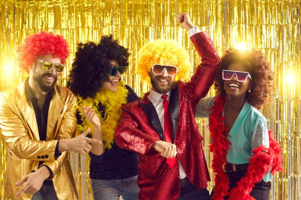 Happy people dancing gangnam style on stage with shiny golden background. Group of friends disguised in boas, glasses, sequin jackets and funny silly curly wigs having fun at night club disco party