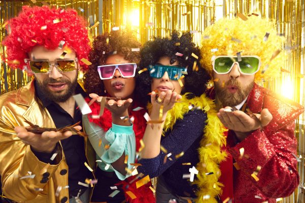 People dressed in boas, party glasses and curly Afro wigs celebrating happy event at disco night club and blowing a kiss with shiny sparkling golden confetti to share positive festive mood with you