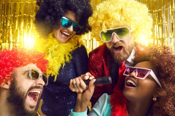 Happy people dressed in funny fancy costumes having fun at music show or karaoke disco party. Group of male and female friends with mikes singing, shouting and dancing on stage with gold background