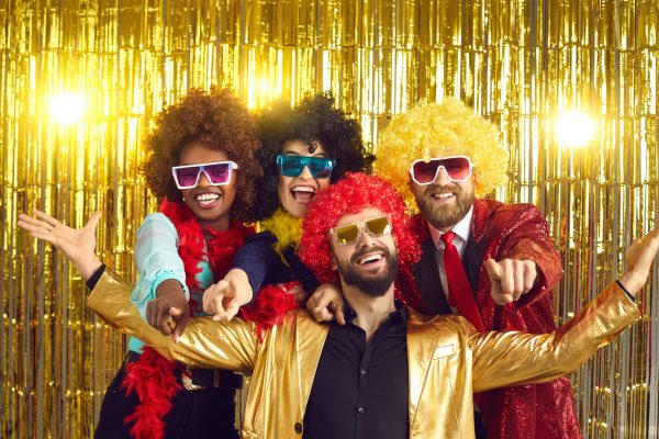 Happy friends point fingers at camera inviting you to join 70s style fancy dress fun groovy disco party. Funny photobooth group portrait of excited young people at New Year Eve or birthday celebration