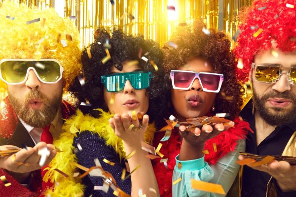 Happy merry people dressed in Afro wigs and glasses celebrating Christmas, New Year, birthday or Xmas Eve at disco night club party sharing groovy festive mood blowing you shiny golden confetti kiss